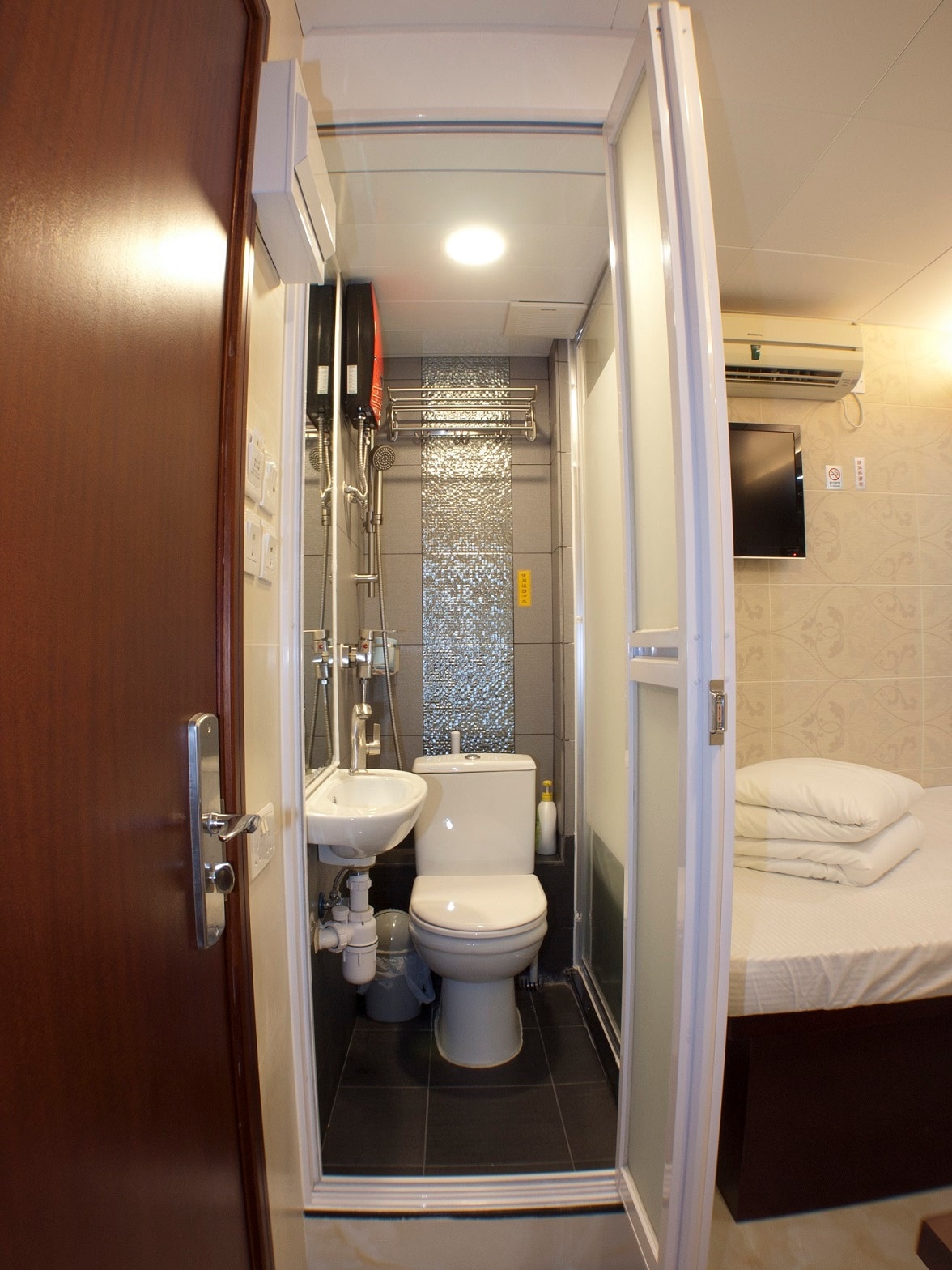 A compact bathroom is visible, featuring a toilet, sink, and shower enclosed in a glass cubicle. Modern fixtures and grey tiles enhance the space. A neatly made bed with fresh linens is located in the adjacent room, suggesting a practical design for efficiency.