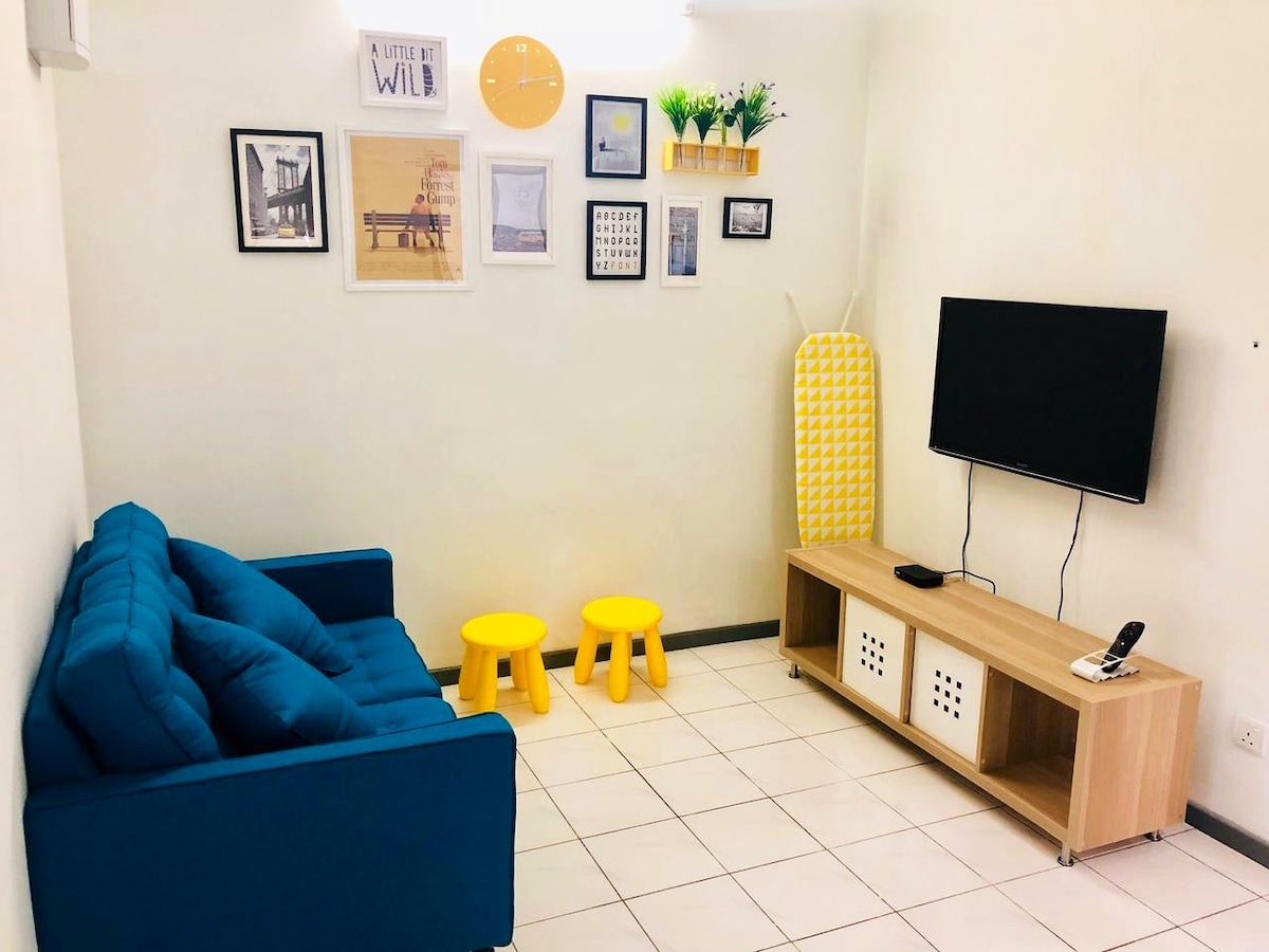 A cozy living room features a blue sofa facing a wooden television unit. Two yellow stools are arranged in front of the unit. The wall displays framed art and decorative shelves, adding a touch of color to the space. A yellow ironing board leans against the wall.