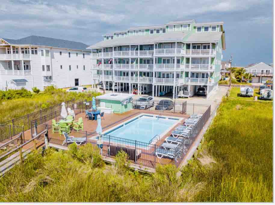 A multi-story building showcases a well-maintained pool area surrounded by lounging chairs. The grassy landscape enhances the outdoor setting, while parking spaces beneath the building are visible. The structure features a light green exterior with large balconies, allowing for outdoor relaxation.