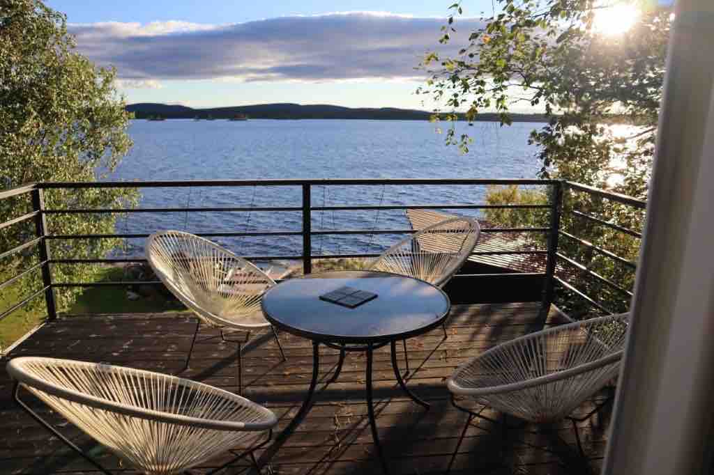 A cozy outdoor terrace is equipped with four circular chairs positioned around a round table, offering views of the serene water and distant hills. Sunlight reflects off the surface, enhancing the peaceful atmosphere of the setting.