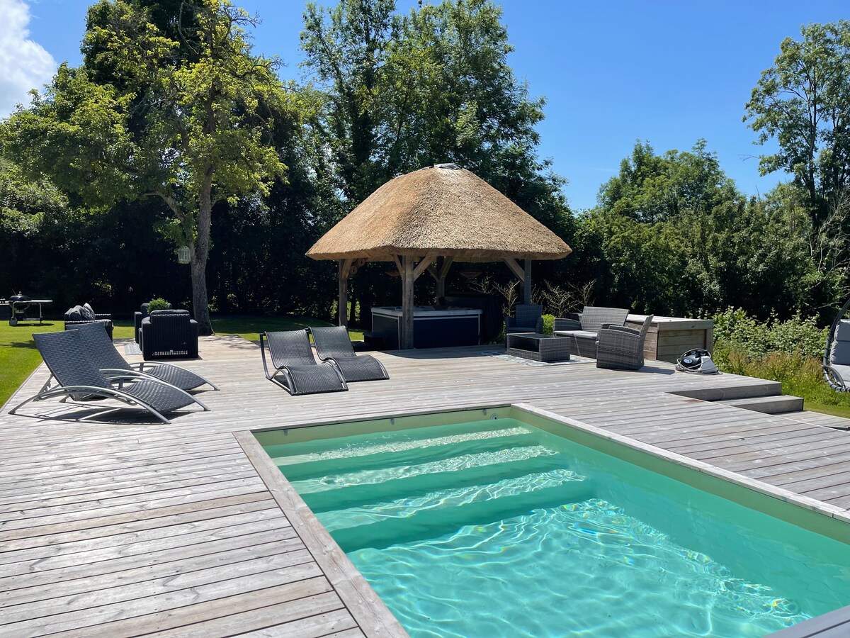 A private, heated pool is bordered by a spacious wooden deck. Relaxing lounge chairs are positioned around the pool, while a thatched-roof gazebo offers shade. Lush greenery surrounds the area, providing a serene environment for guests to unwind.