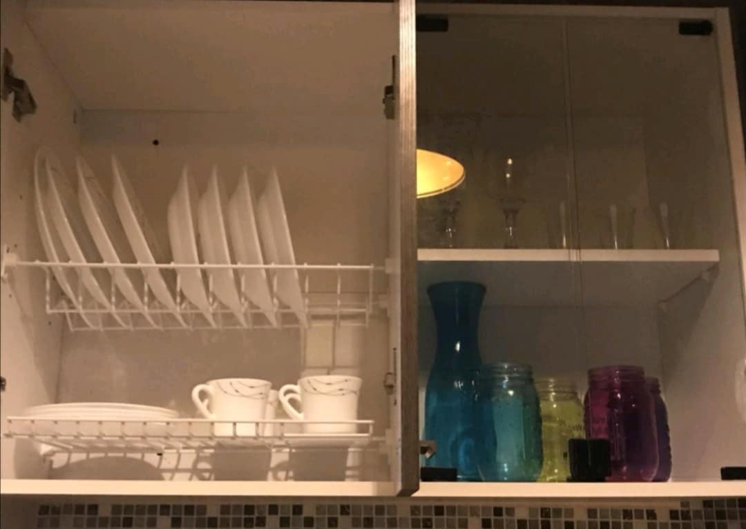 A neatly organized kitchen cabinet is shown, featuring white plates and cups arranged on a drying rack. Colored glass vases and jars in shades of blue, green, and purple are displayed on the shelves, creating a vibrant yet orderly appearance.