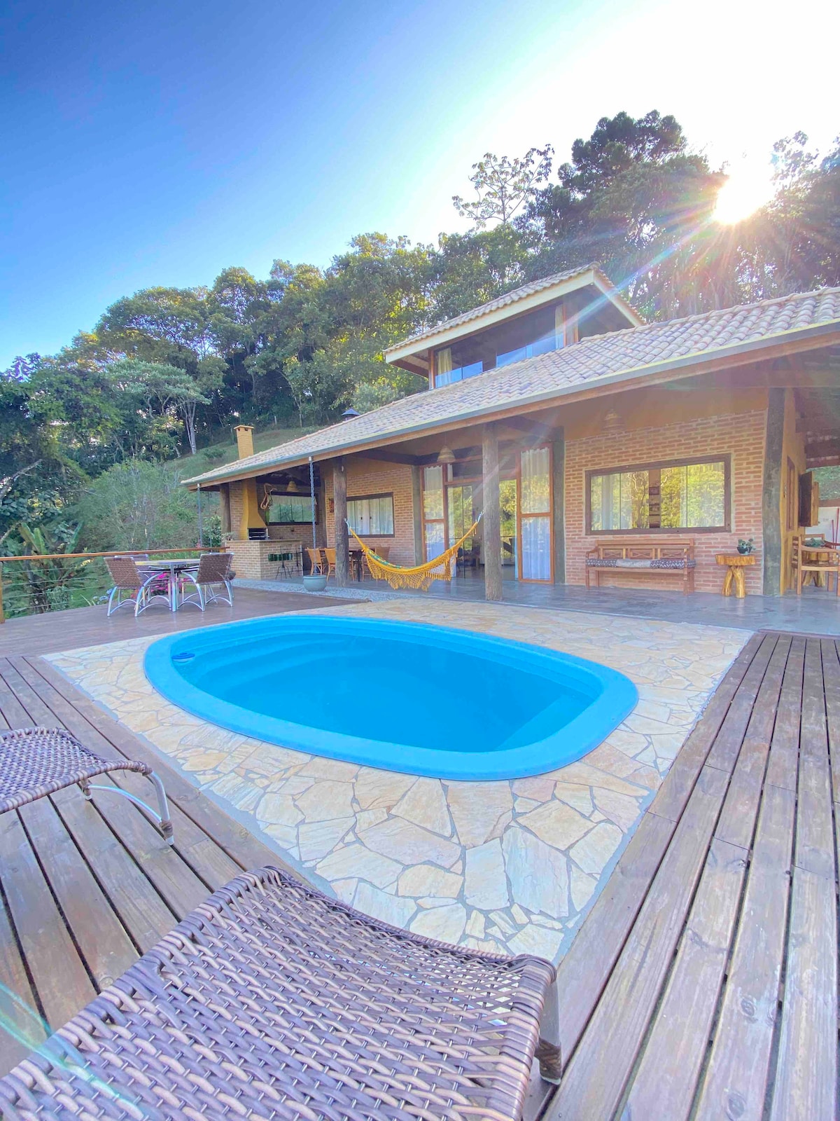 An inviting outdoor area features a refreshing blue pool surrounded by a wooden deck. Comfortable lounge chairs are positioned nearby, with a hammock suspended between two posts. The house, with its warm brick exterior and spacious windows, is set against a backdrop of lush greenery.