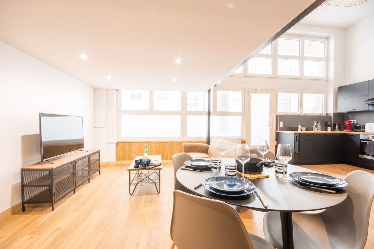 An open living area features a dining table set with plates and glasses, accompanied by a cozy sofa and a TV. Large windows allow natural light to fill the space, highlighting wooden flooring and modern furniture.