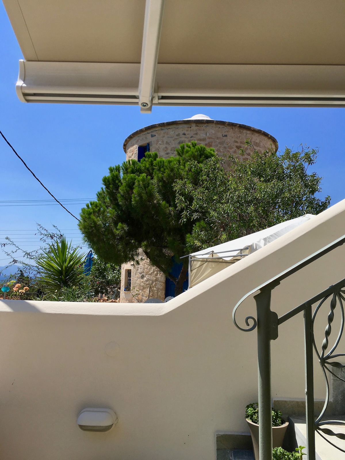 The image captures a view of a stone windmill from a stairway. Lush greenery is visible alongside the structure, with a clear sky above. A shaded terrace area is discernible, enhancing the inviting outdoor space. The scene conveys a charming blend of nature and architecture.