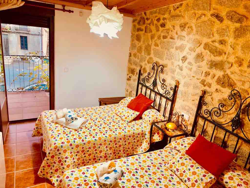 The room features two twin beds, each adorned with floral-patterned bedding and complemented by red accent pillows. A warm light fixture hangs above, while natural light enters through a window overlooking a quiet terrace. Exposed stone walls contribute to a charming atmosphere.