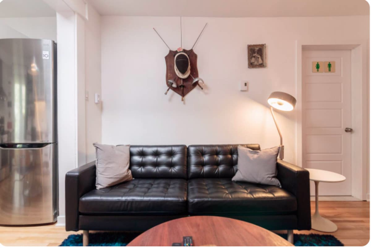 A comfortable black leather sofa is adorned with two decorative pillows. A round coffee table is placed in front of the sofa, while a modern floor lamp provides illumination. A mounted wall decor piece is visible above the sofa, and a metal fridge stands to the left.