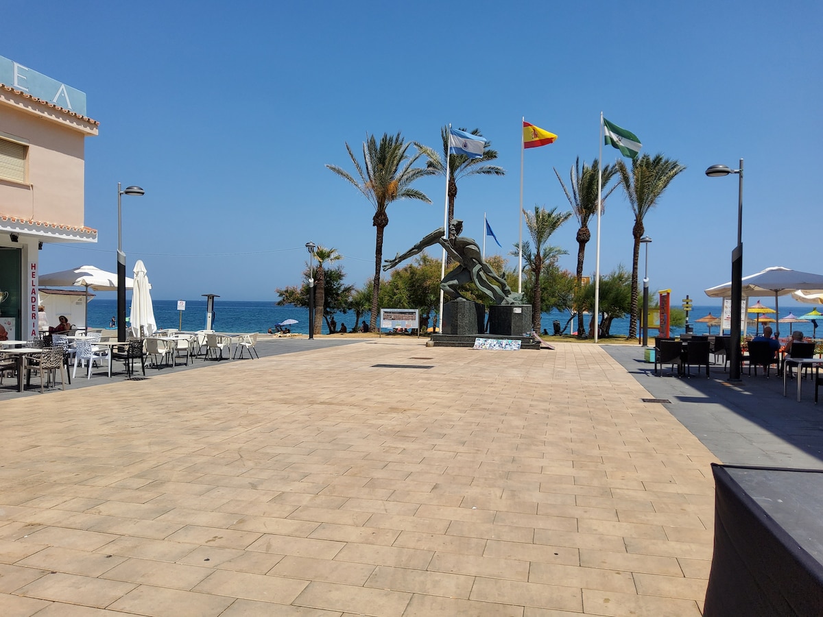 An open plaza is visible, lined with palm trees and umbrellas providing shade. A sculpture of a turtle is positioned at the center, with flags displayed nearby. The beach and ocean can be seen in the background under clear blue skies.