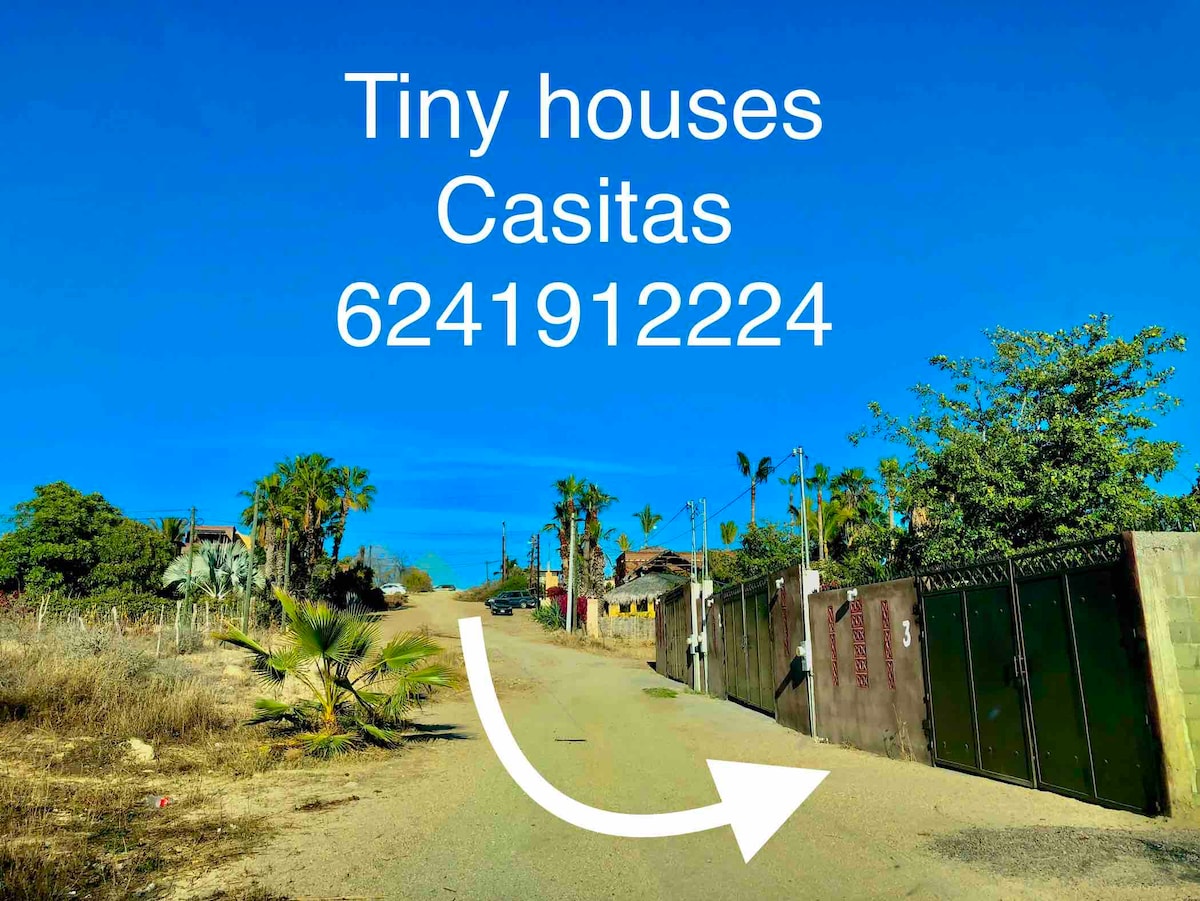 An inviting view of a dirt road leads to a gated entry featuring green gates adorned with numbers. Lush greenery and palm trees border the pathway, with a clear blue sky overhead, providing a sense of tranquility and approachability.