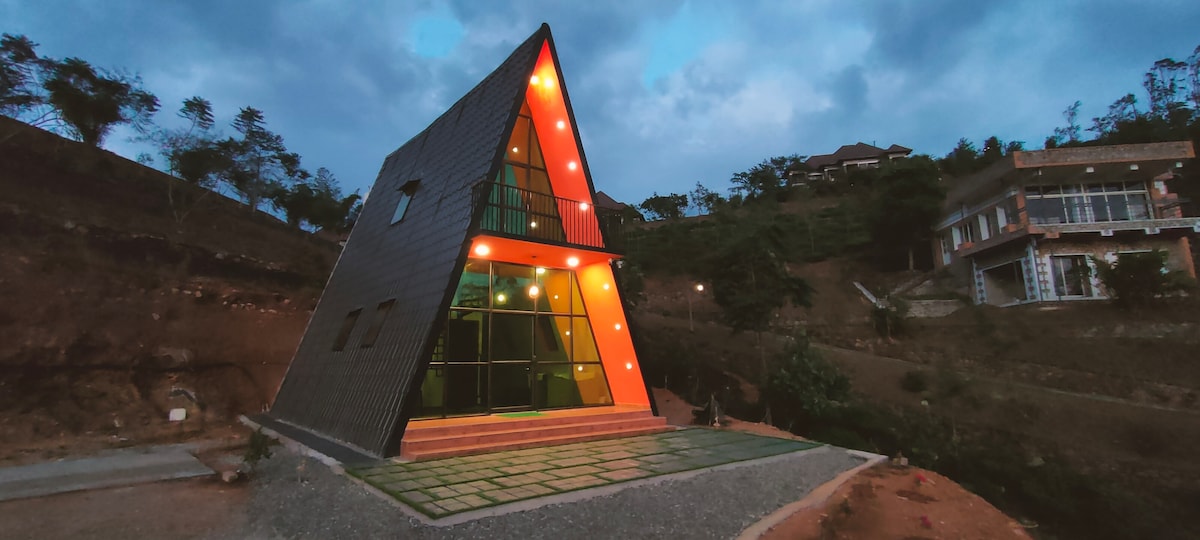 A modern A-frame structure is positioned on a hillside, featuring large glass windows and a striking orange glow from the exterior lighting. A well-maintained path of stone pavers leads to the entrance, surrounded by natural landscaping and distant greenery.