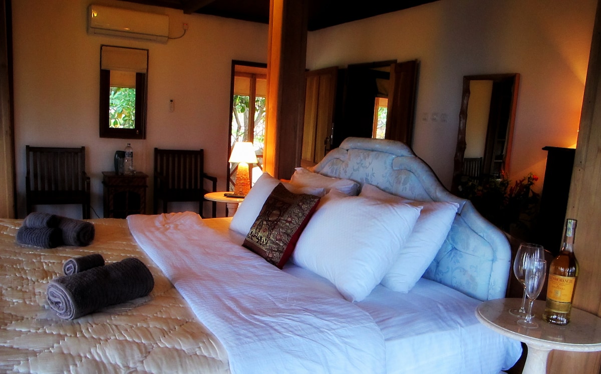 A spacious bedroom features a king size bed adorned with plush pillows and a decorative throw. Natural light enters through a nearby window, illuminating the room's wooden accents and elegant furniture, including two chairs and a side table with a bottle and glasses.