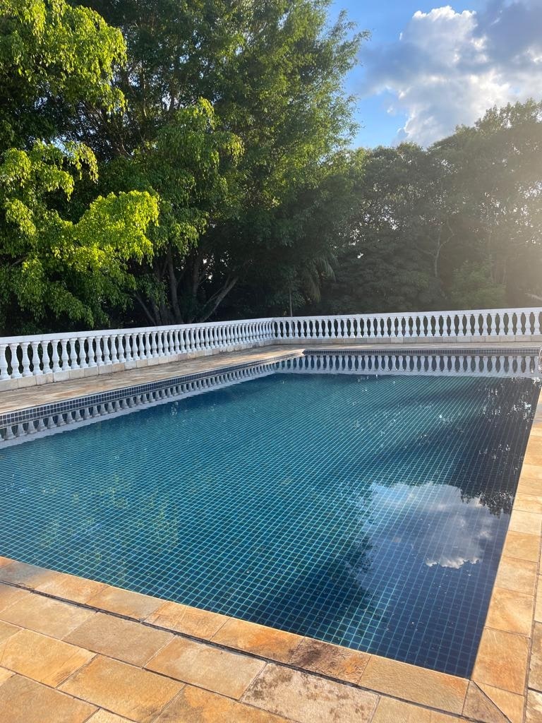 A clear swimming pool is surrounded by a stone patio and embellished with decorative railings. Lush greenery provides a natural backdrop, complementing the bright sky. Sunlight reflects on the water's surface, highlighting the pool's inviting appearance.