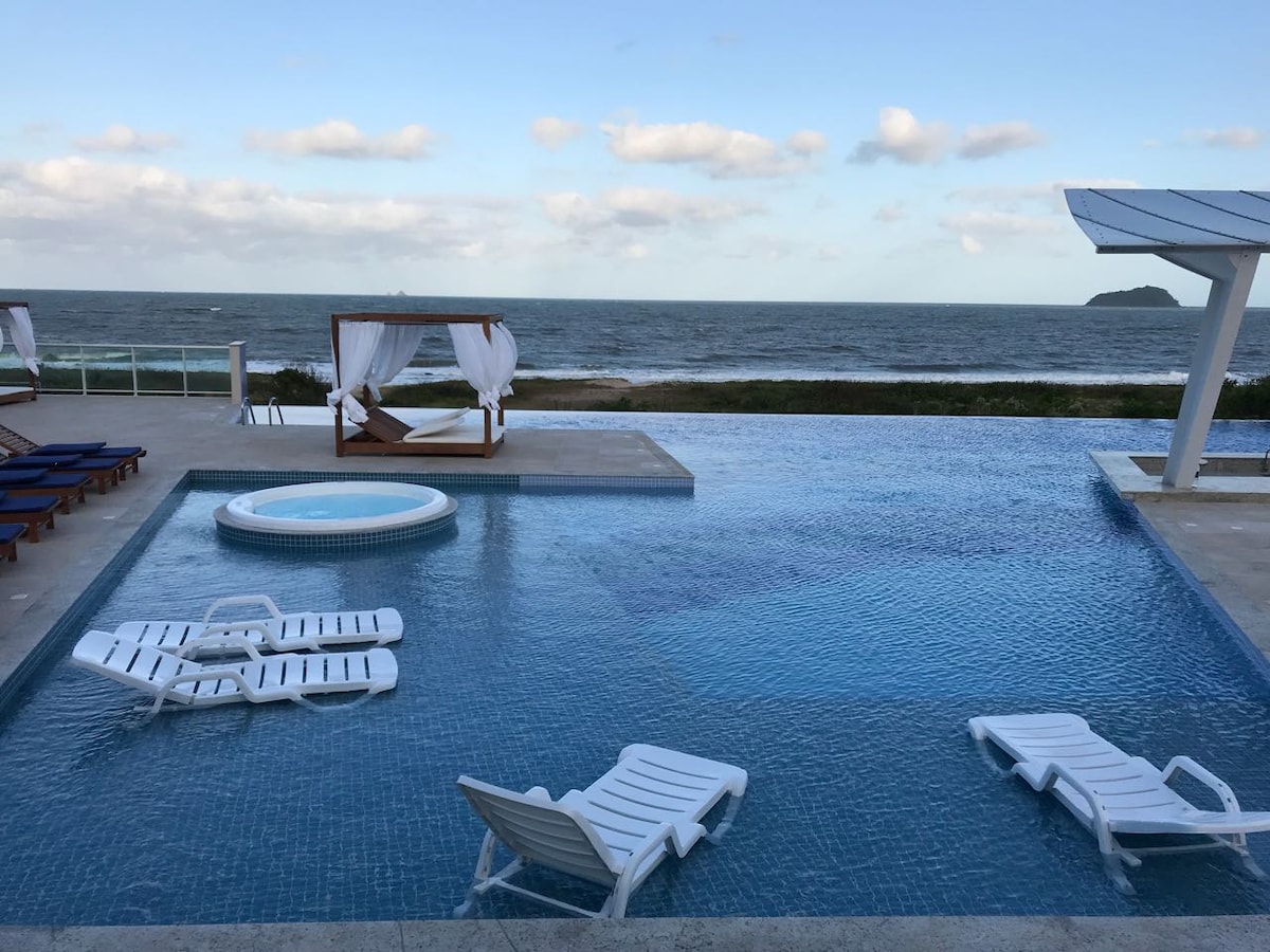 An outdoor infinity pool overlooks the ocean, featuring a small jacuzzi adjacent to a spacious lounging area with cabanas. Multiple white lounge chairs surround the pool, providing comfortable spaces for relaxation. The expansive view of the sea is framed by a clear sky.