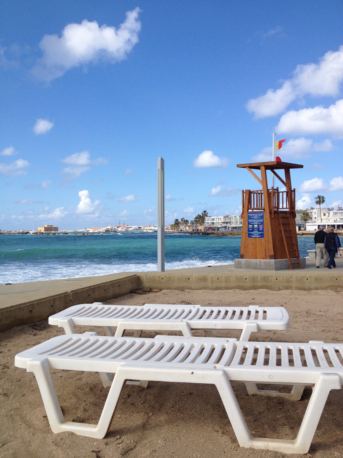 Two white sun loungers are placed on a sandy beach, facing the calm blue sea. A lifeguard tower stands nearby, equipped with a red flag. The shoreline features a backdrop of coastal buildings and a clear blue sky with scattered clouds.