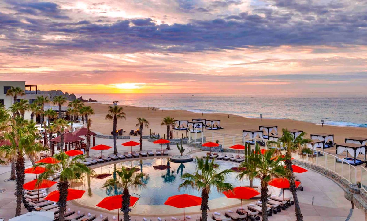 A resort-style pool is surrounded by lounge chairs and vibrant red umbrellas. Palm trees provide shade, while the beach and ocean are visible in the background. A sunset casts warm tones across the sky, enhancing the serene atmosphere of the space.