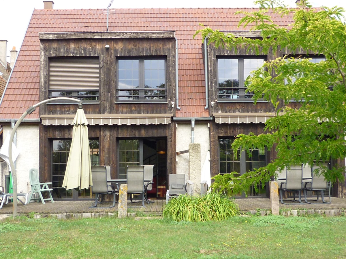 The exterior features a two-story building constructed with wooden elements and a red tiled roof. A spacious patio area is presented, furnished with several chairs and a table, along with a large umbrella for shade. Lush greenery is visible in the foreground.
