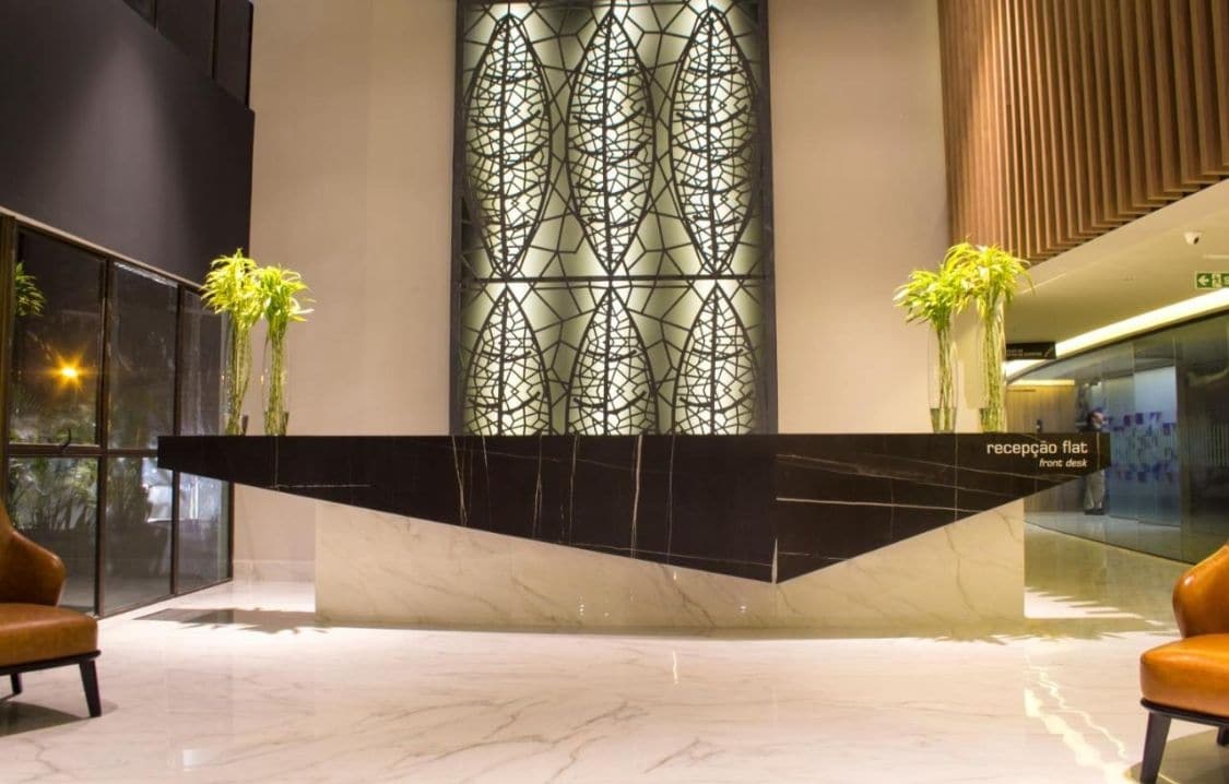 The lobby features a sleek reception desk made of dark stone, accented by contemporary design elements. Decorative plants are placed on either side, adding a touch of greenery. A large glass wall allows for natural light, complemented by stylish lighting fixtures overhead.