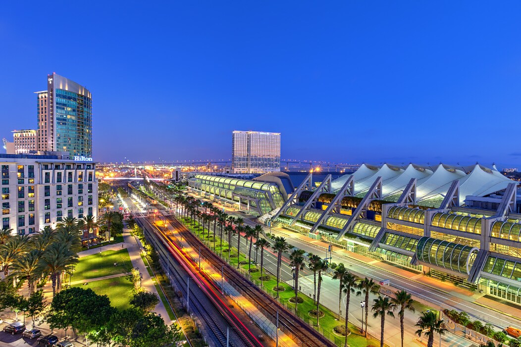 A vibrant evening view of downtown San Diego showcases the Convention Center, adorned with distinctive architectural features and large glass facades. Palm trees line the street, while city lights twinkle in the background. A train can be seen passing through, adding movement to the scene.