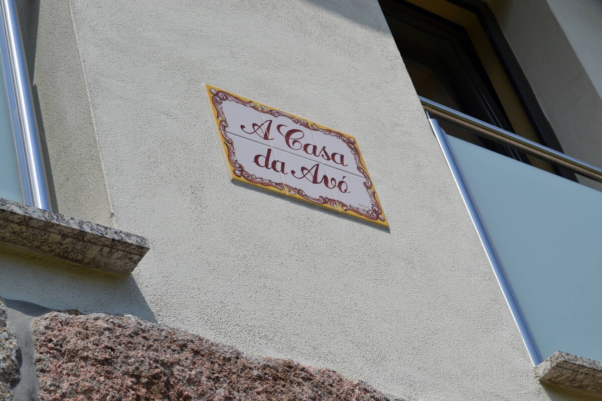 The exterior of the property features a decorative sign that reads 'A Casa da Avó,' positioned above a stone façade. A balcony with glass railings can be seen, allowing for natural light to enhance the area.
