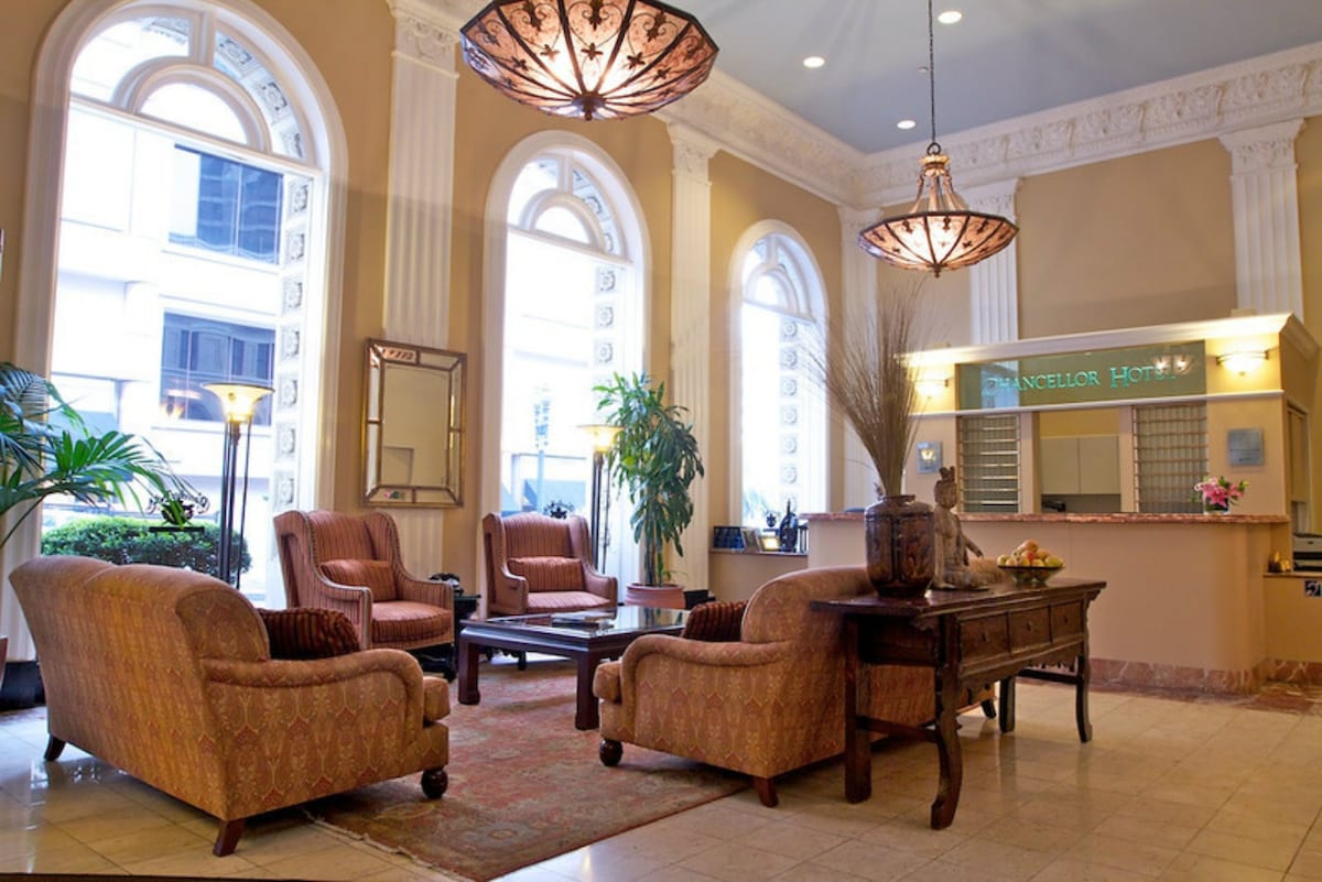 A welcoming lobby area features elegant furnishings, including two patterned armchairs and a matching sofa. The space is highlighted by large windows allowing natural light and adorned with decorative plants. A reception desk is visible in the background, complemented by ambient lighting from stylish fixtures.