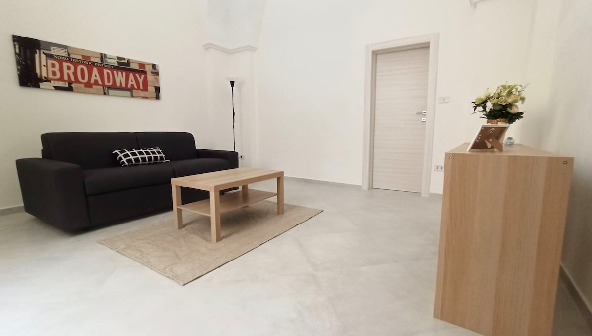 A living area is presented with a dark sofa positioned opposite a wooden coffee table. A light area rug lies beneath the table. A decorative sign stating 'BROADWAY' hangs on the wall. A minimalist cabinet can be seen to the right, with a vase of flowers placed on top.