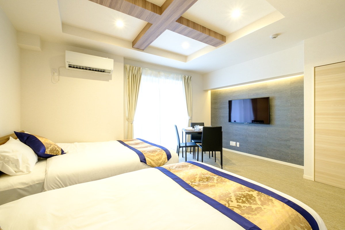 A well-lit room features two spacious beds with decorative throws, positioned beside a window with sheer curtains. A small dining table and chairs are visible, along with a mounted flat-screen television against a textured wall.