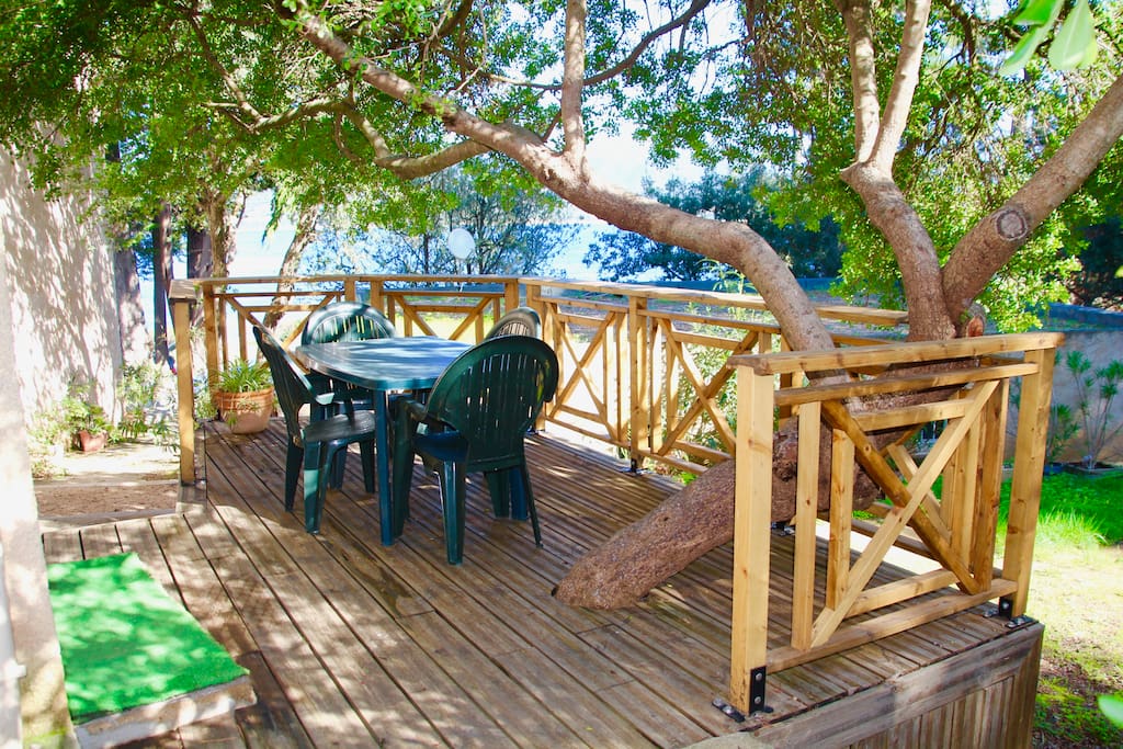 La terrasse en bois de l'appartement construite autour d'un superbe lentisque. Pour une détente et des repas ombragés uniques. Au fond, la vue mer est très réduite mais elle ne se trouve qu'à 40 m.