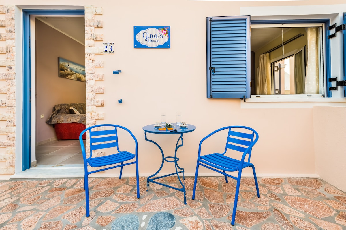 A charming exterior features a light-colored wall with a welcoming sign. Two bright blue chairs are set around a small table, positioned on a stone patio. A window with blue shutters offers a peek inside, while a partial view of a cozy bed can be seen through the glass.