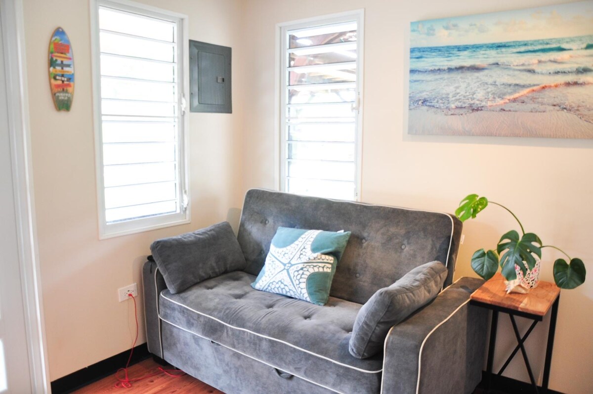 A comfortable sofa with a decorative cushion is positioned near bright windows, allowing natural light to illuminate the space. A small table holds a potted plant, and a beach-themed artwork adds a calming touch to the wall.