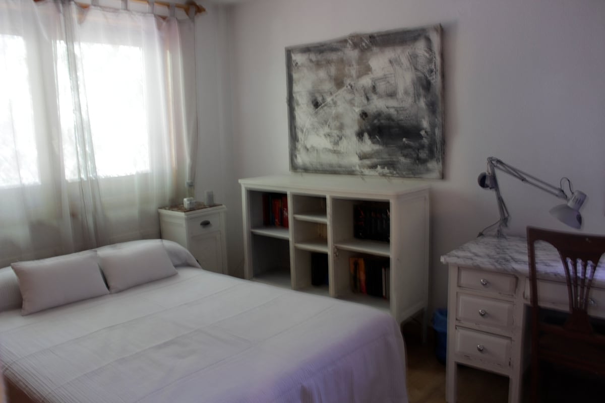 A well-lit bedroom features a spacious bed adorned with simple white linens. A large piece of abstract artwork hangs on the wall above a stylish bookshelf. A desk with a lamp occupies one corner, complemented by a wooden chair.