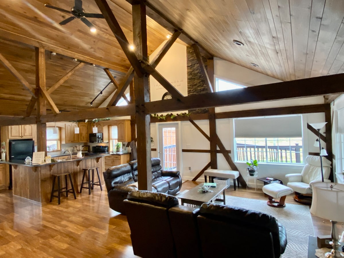 An open-concept living area features wooden beams and a combination of rustic and modern designs. Leather seating is arranged thoughtfully, with a coffee table at the center. A spacious kitchen is visible in the background, complemented by large windows allowing natural light to fill the space.