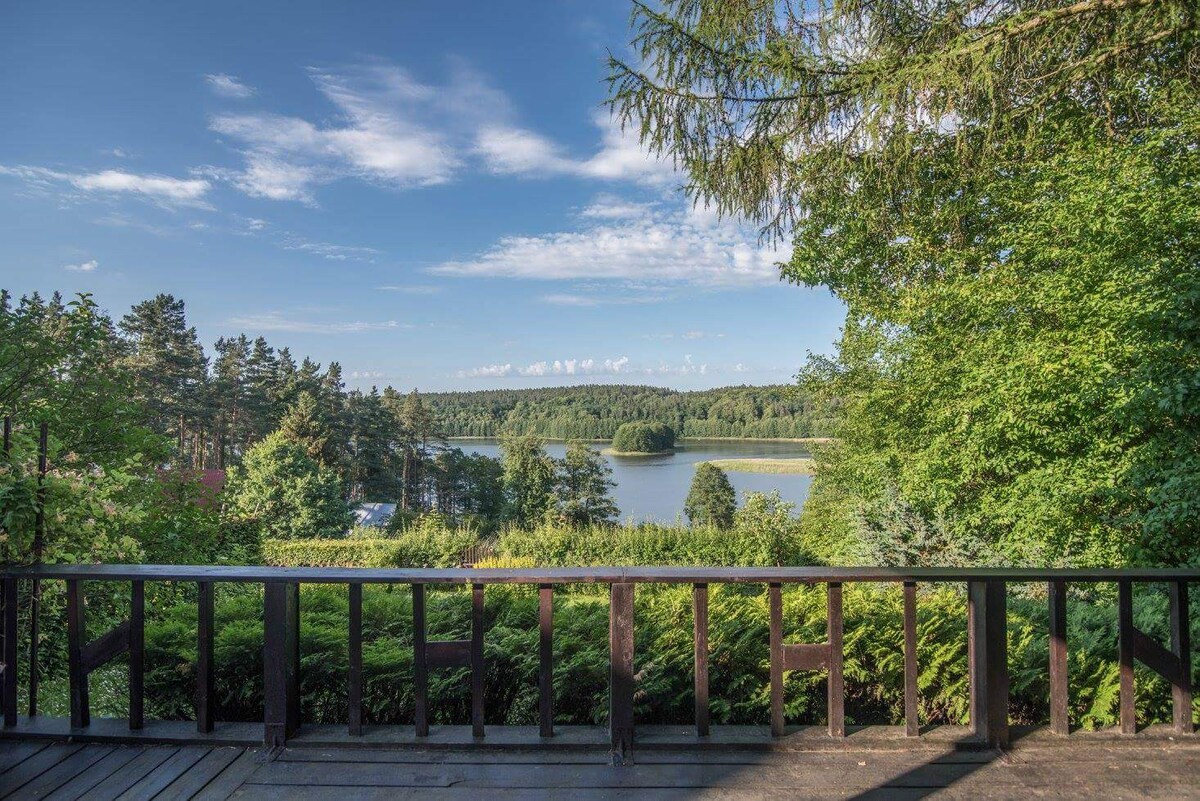 A view from a wooden terrace overlooks a serene lake surrounded by lush greenery. The calm water reflects the blue sky, punctuated by scattered clouds and framed by trees, creating a peaceful getaway scene.