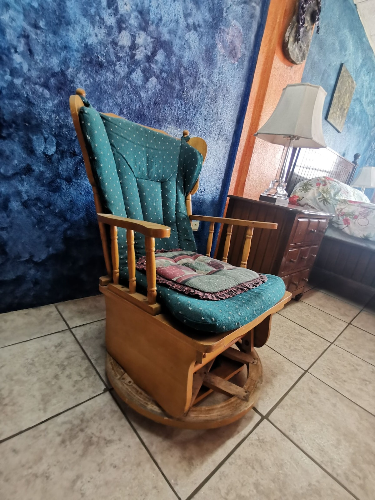 A wooden rocking chair with a turquoise cushioned seat is positioned against a textured blue wall. A quilted blanket is draped across the backrest, while nearby furniture, including a bedside lamp and wooden dresser, is partially visible.