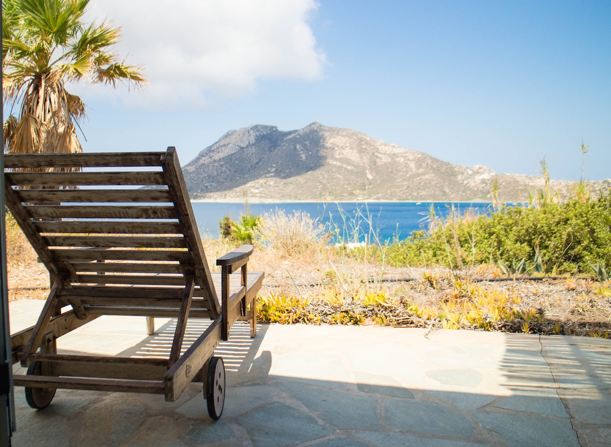 A wooden lounge chair is positioned on a stone patio, facing a stunning view of the sea and mountains. Warm sunlight illuminates the scene, while vibrant greenery surrounds the area, creating a serene outdoor space ideal for relaxation.