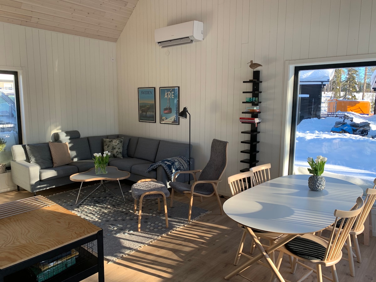 A spacious living area features a modern sectional sofa, accent chairs, and a light wood dining table surrounded by chairs. Natural light enters through large windows, illuminating the room. Decorative plants and wall art add subtle decoration, enhancing the inviting ambiance.