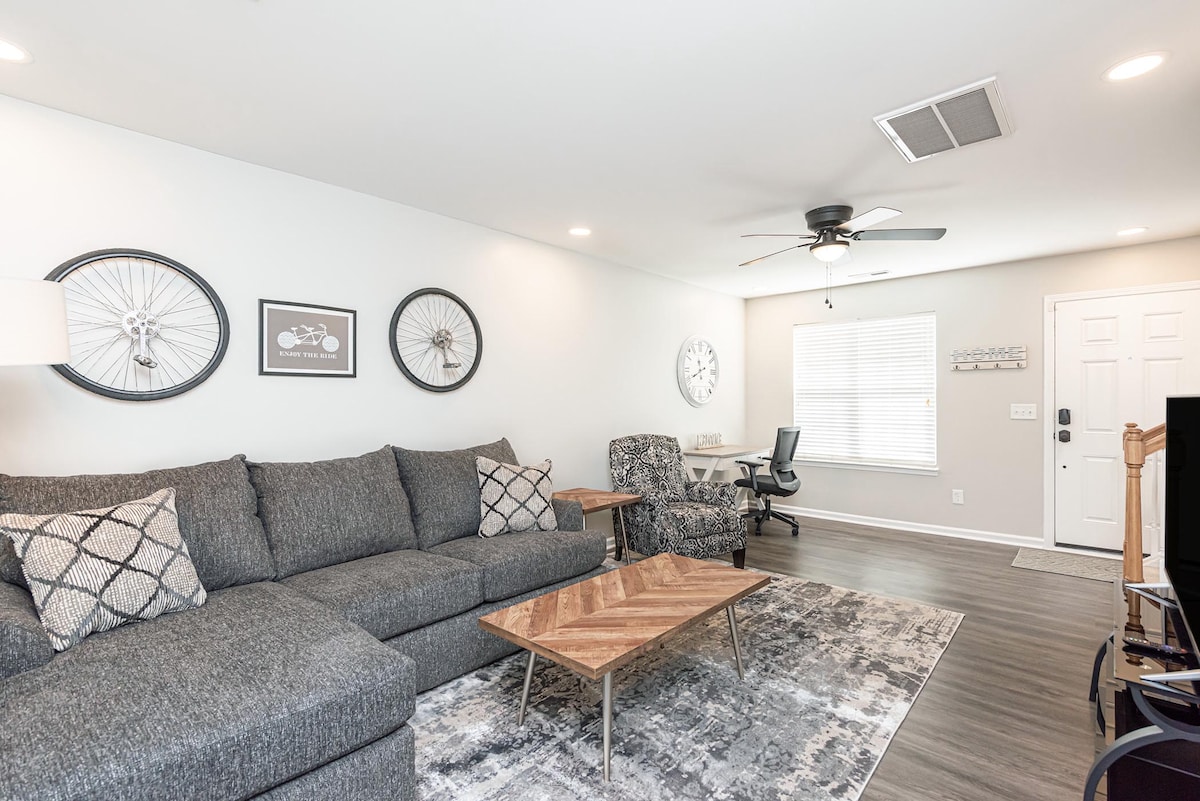 A spacious living area features a large, comfortable sectional sofa with decorative pillows, complemented by a stylish wooden coffee table. Artwork, including bicycle wheels, adorns the walls, and natural light brightens the room through a window, with a ceiling fan providing added comfort.