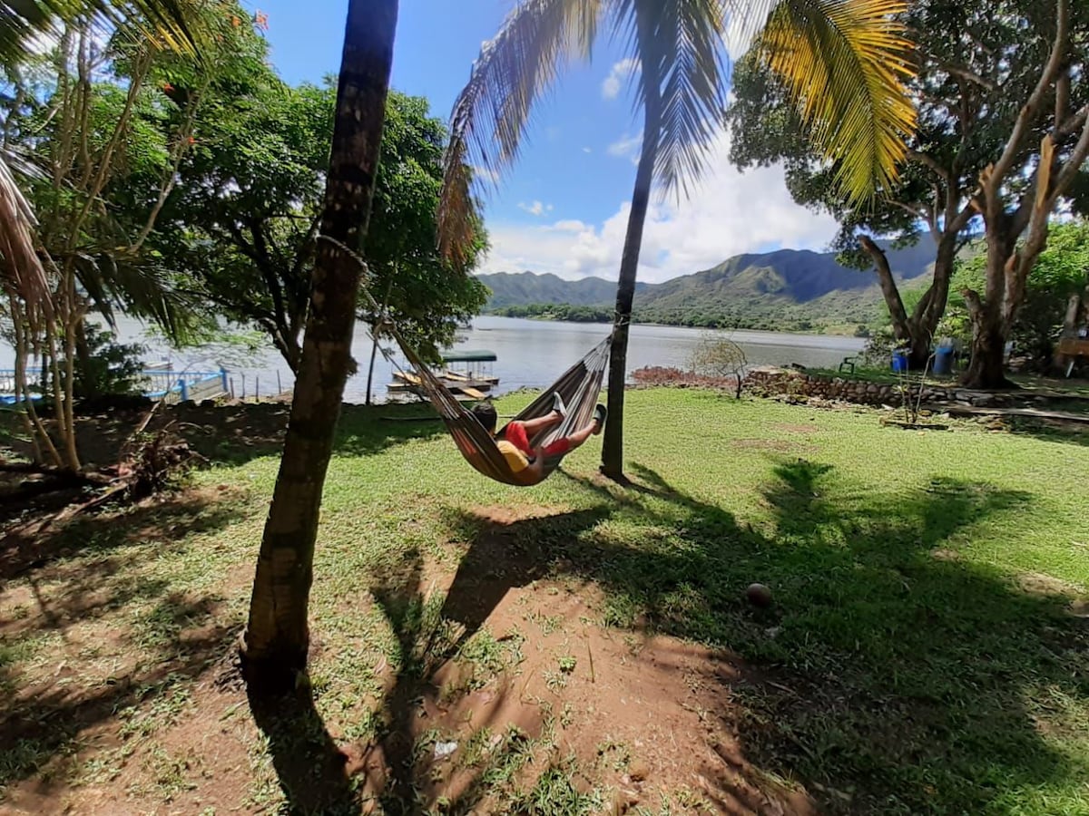 A hammock is suspended between two palm trees in a grassy area, overlooking a serene lake and surrounded by lush greenery. Gentle sunlight casts dappled shadows on the ground, while distant mountains are visible in the background.