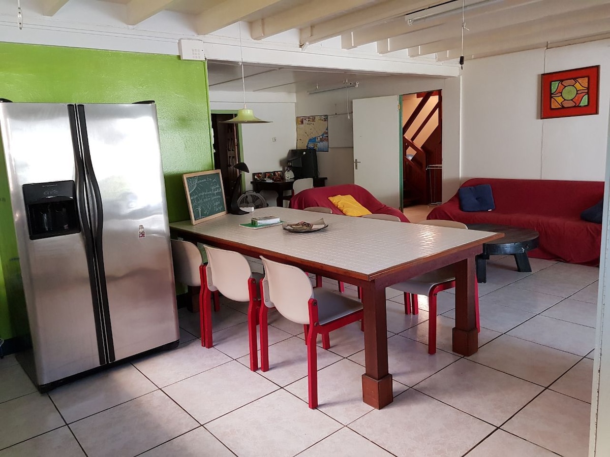 A spacious dining area features a large table surrounded by bright chairs, inviting communal meals. The kitchen area includes a stainless steel refrigerator and a green accent wall, while a cozy seating area is present with various seating options, enhancing the open layout.