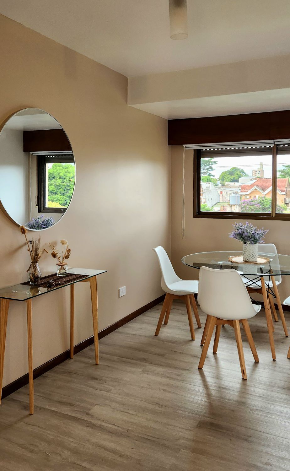 A dining area features a glass table surrounded by four white chairs, with a decorative round mirror mounted on the wall. Natural light enters through two windows, offering a view of the neighborhood. Subtle floral arrangements add a touch of decor.