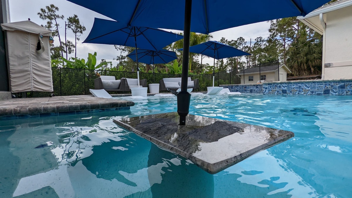 A cocktail-style saltwater pool is depicted, featuring in-water lounges and umbrellas that provide shade. A submerged table is visible in the center, enhancing the pool area. Surrounding foliage suggests a serene outdoor environment, complementing the inviting blue water.