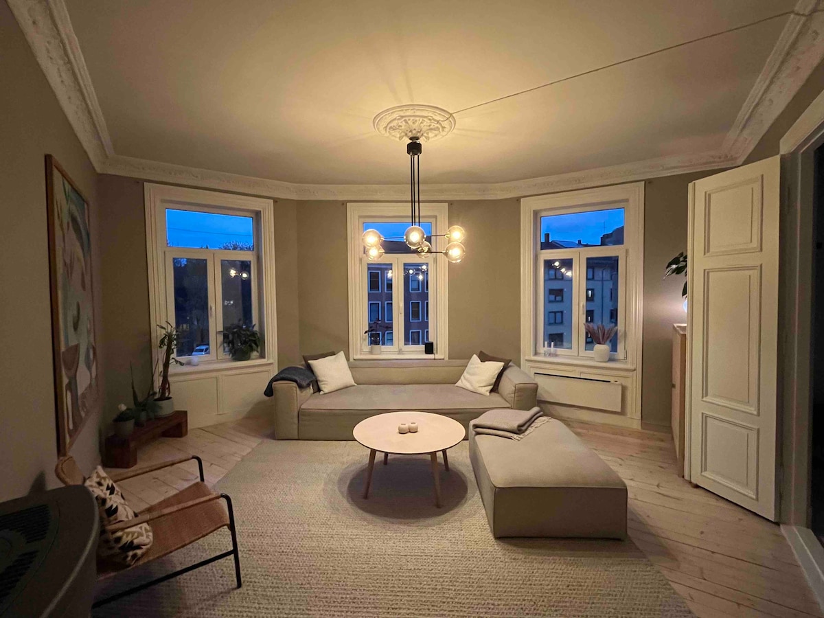 A spacious living area is presented, featuring a large sectional sofa and a round coffee table at its center. Multiple windows allow for natural light, accented by an elegant light fixture overhead. Subtle decor elements are visible, enhancing the overall ambiance.