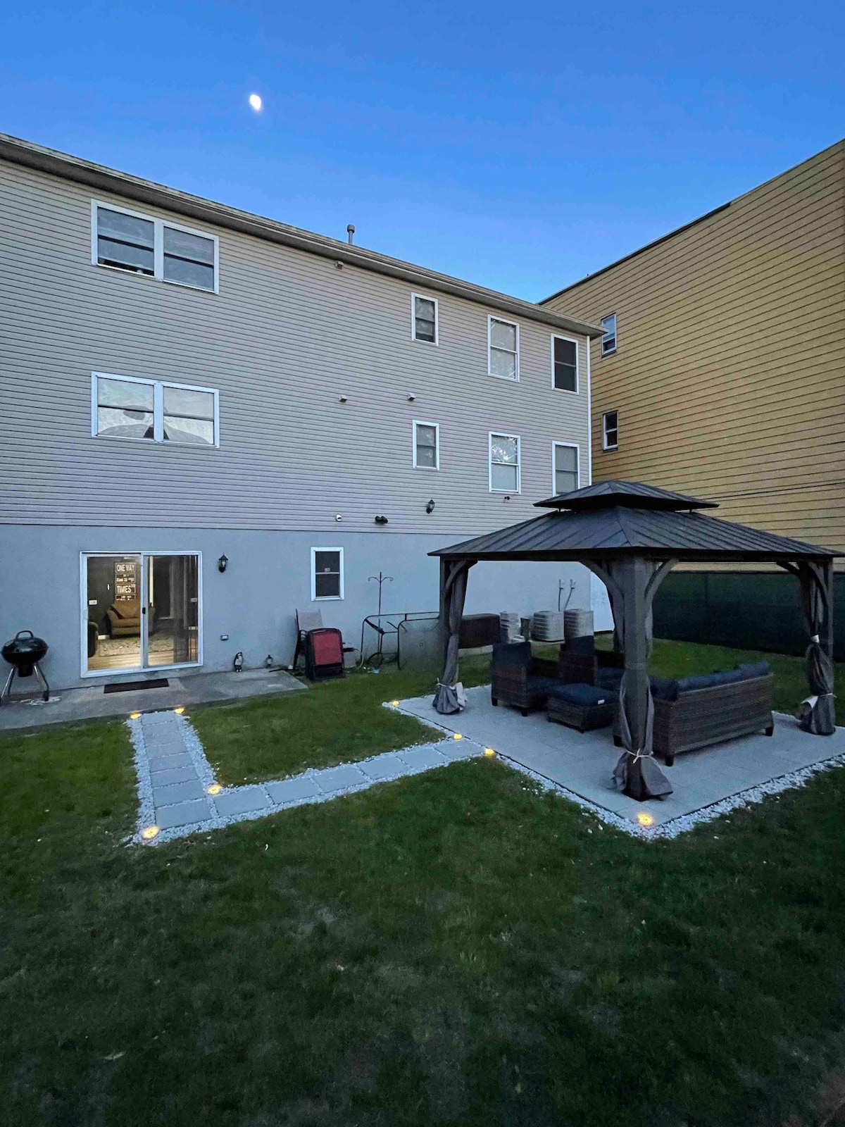 A well-maintained backyard features a patio area shaded by a gazebo, furnished with dark lounge seating. Pathways lined with soft lighting create an inviting atmosphere. The exterior of a multi-family home can be seen in the background, highlighting accessible outdoor space.