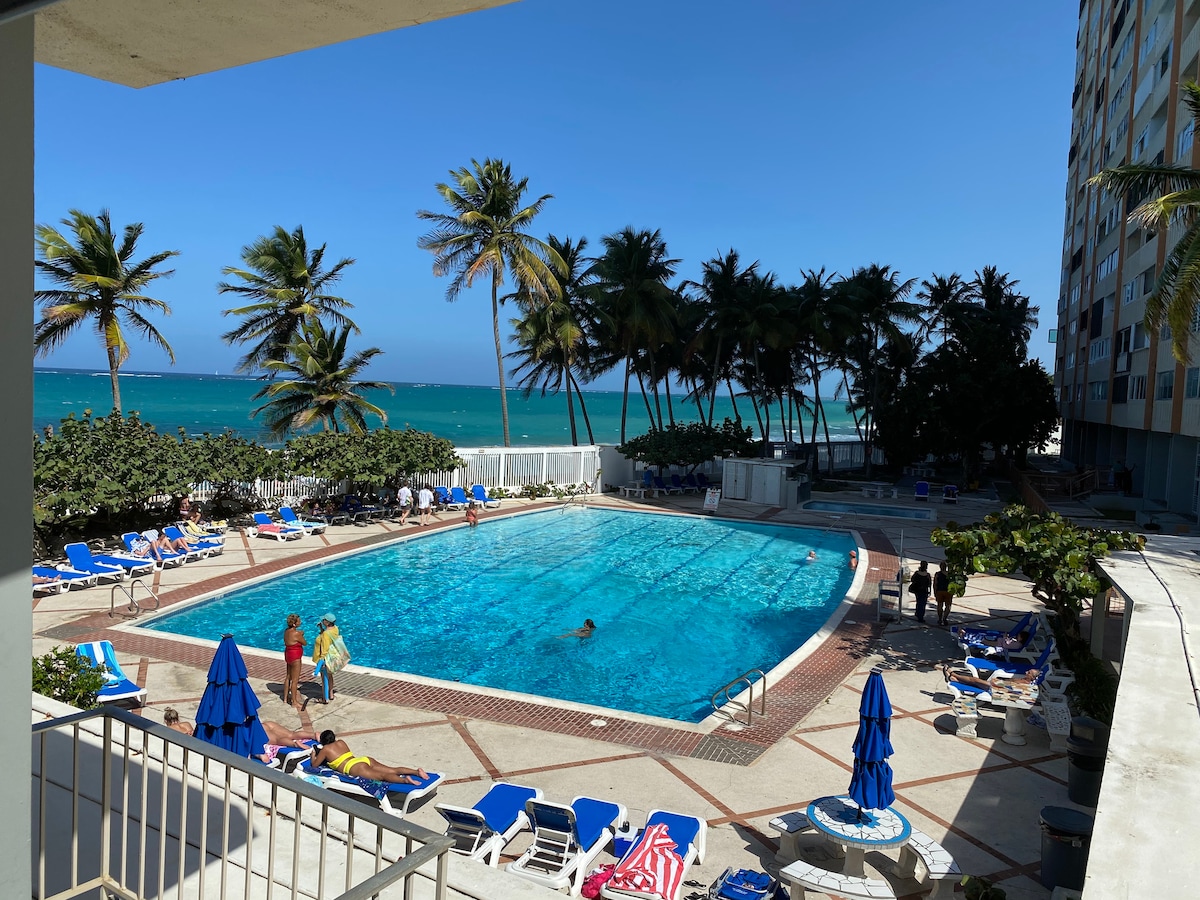 An expansive swimming pool is framed by lounge chairs and umbrellas, inviting relaxation under the sun. Palm trees sway in the gentle breeze, and the ocean is visible in the background. People enjoy the cool water, adding a lively atmosphere to the serene setting.