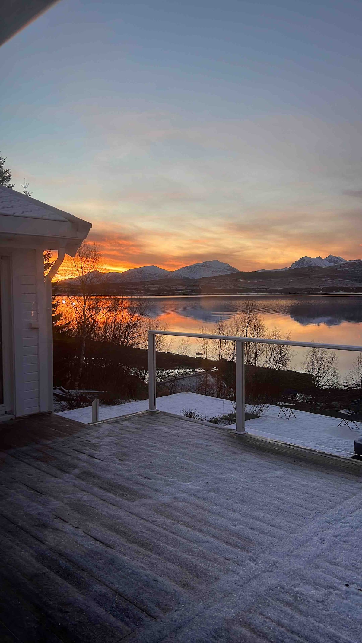 A scenic view from a deck overlooks a calm lake reflecting the colors of a sunrise. Snow-covered grounds and trees surround the area, while distant mountains provide a picturesque backdrop against the warm hues of the morning sky.