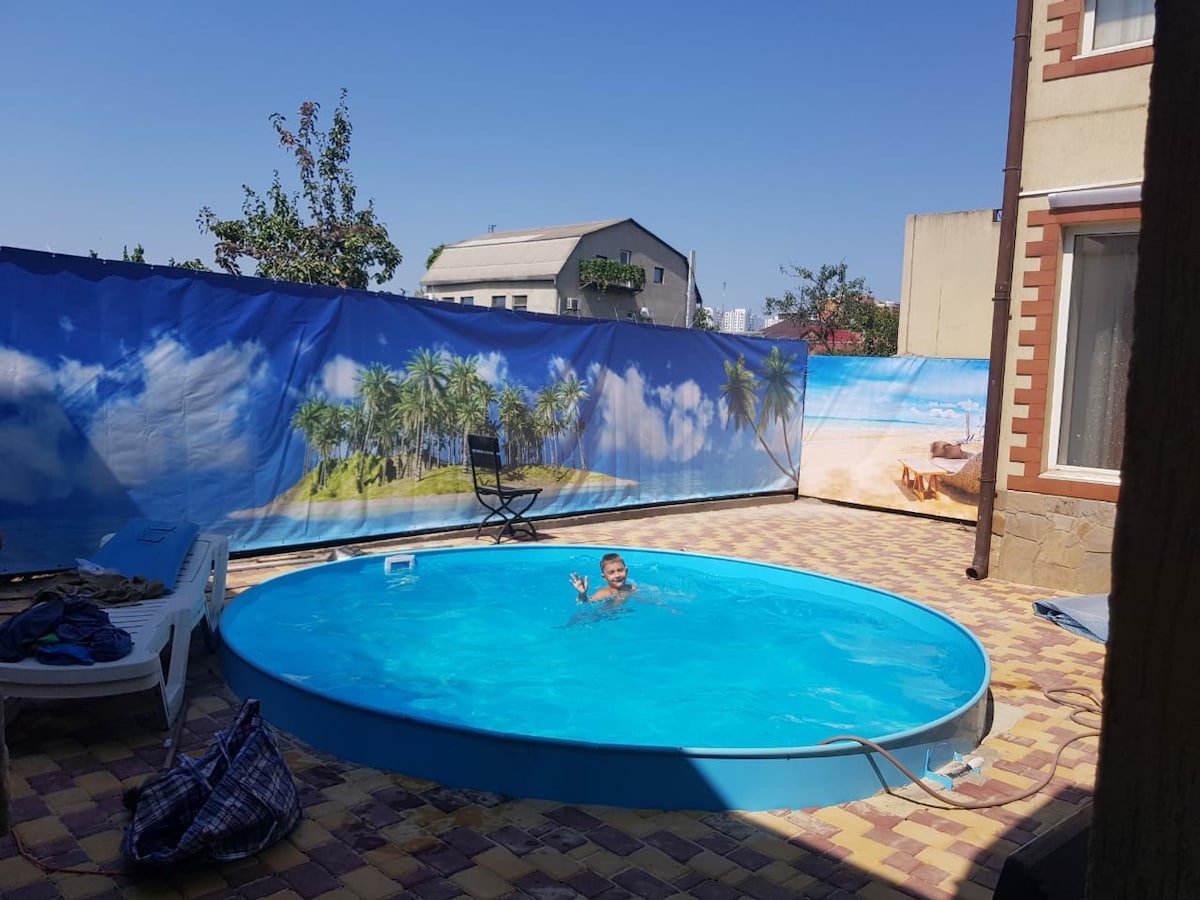 A circular outdoor pool is surrounded by colorful paving stones. A blue sky is visible overhead, and palm tree artwork is displayed on a barrier around the pool area. A single person is enjoying the pool, adding a sense of leisure to the setting.