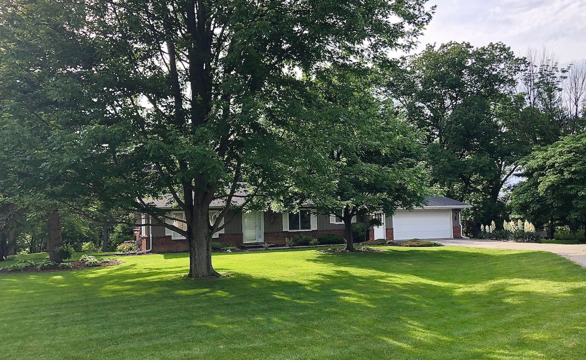 A single-story brick home is framed by mature trees, offering shade to the lush green lawn. A paved driveway leads to a garage on the right, while flower beds border the front, adding a touch of color to the landscape.