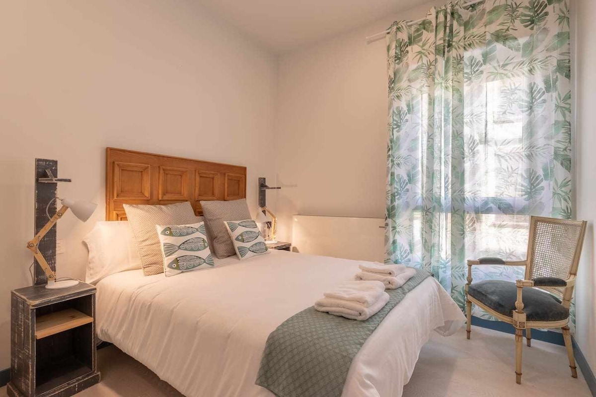A serene bedroom features a spacious bed adorned with soft linens and decorative pillows. Natural light filters through sheer curtains with a floral pattern. A cozy chair and bedside tables with stylish lamps complete the inviting space.