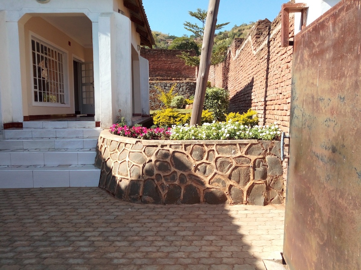 A stone-bordered flower bed displays vibrant flowers in various colors, set against a textured wall. The pathway, made of paved stones, leads to a covered entryway. Lush greenery is visible in the background, complementing the inviting exterior of the guesthouse.