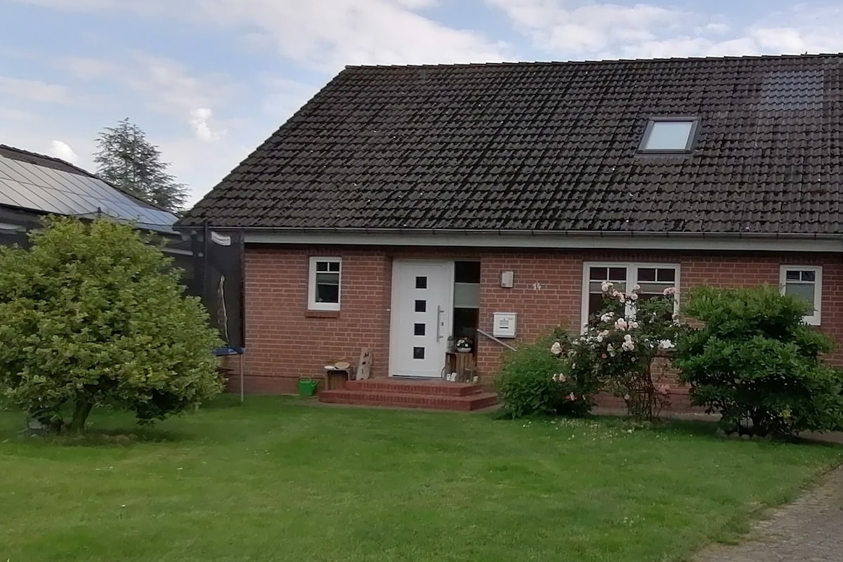 A red-brick house features a sloped roof and a prominent front entrance with a white door. Surrounding greenery includes a well-maintained lawn and shrubs, contributing to a serene outdoor space. A small porch with steps leads to the entrance, enhancing accessibility.