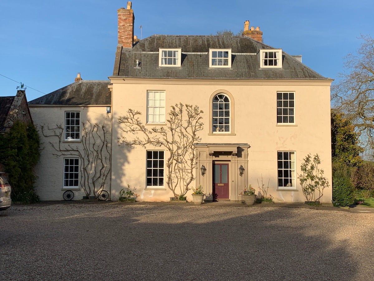 The elegant façade of a two-story grade II listed building is showcased, featuring symmetrical windows and a prominent arched entryway. Climbing plants adorn the walls, and a gravel driveway leads to the entrance, surrounded by manicured greenery.
